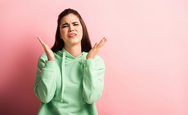 Upset girl looking at camera while standing with open arms pink background